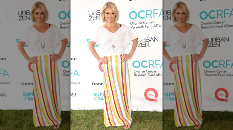 Kelly Ripa posing in a white T-shirt, black bra, and multicolored striped skirt