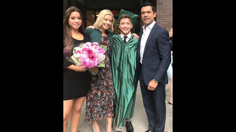 Kelly Ripa wearing a black floral-print dress and green cardigan and posing with her children Lola Consuelos and Joaquin Consuelos and husband Mark Consuelos
