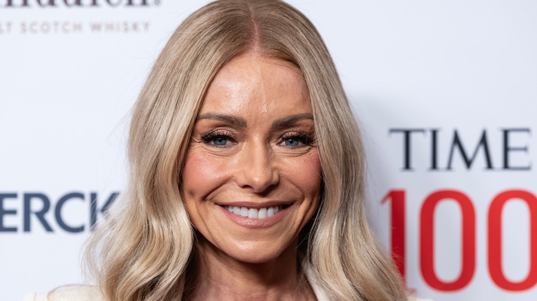 Kelly Ripa smiling in a red dress while wearing a white sweater at the Time 100 gala.