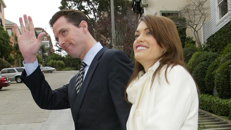 Gavin Newsom waving as he and Kimberly Guilfoyle walk down the street