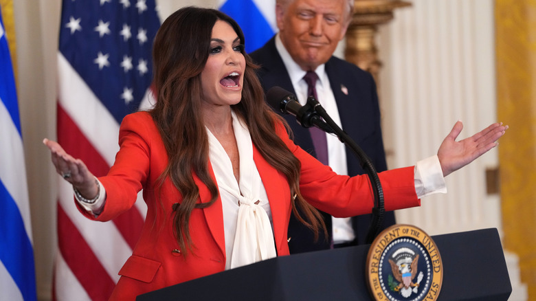 Kimberly Guilfoyle yelling at a podium in a red suit with Donald Trump standing behind her smiling