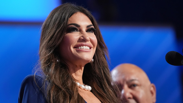 Kimberly Guilfoyle smiling broadly at a podium in a blue dress and pearls