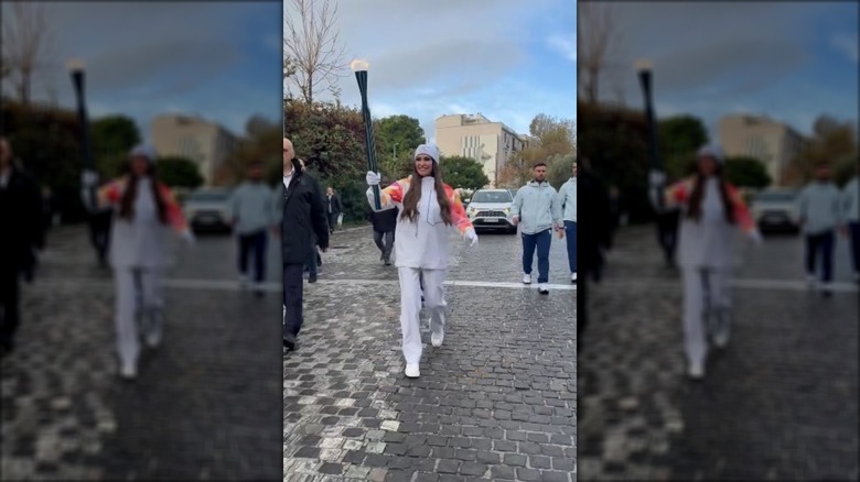 Kimberly Guilfoyle in all-white winter outfit, raising up the Olympic torch on a cobblestone path.
