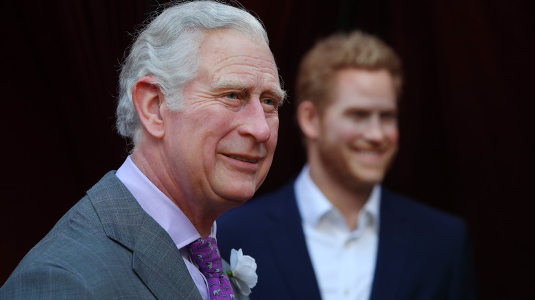 King Charles smiling with a blurry Prince Harry in the background