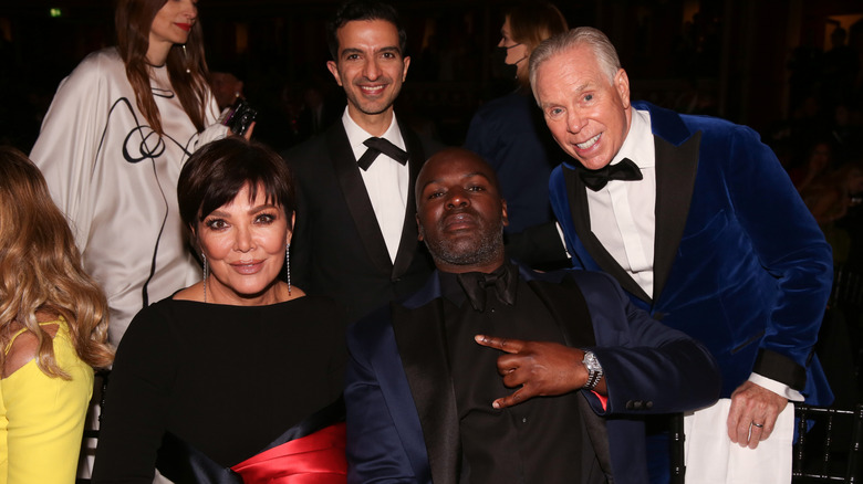 Kris Jenner seated in a black dress beside Corey Gamble as Imran Amed and Tommy Hilfiger pose behind them.