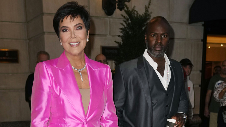 Kris Jenner wears a pink satin suit and smiles beside Corey Gamble, who wears a black satin suit and frowns.