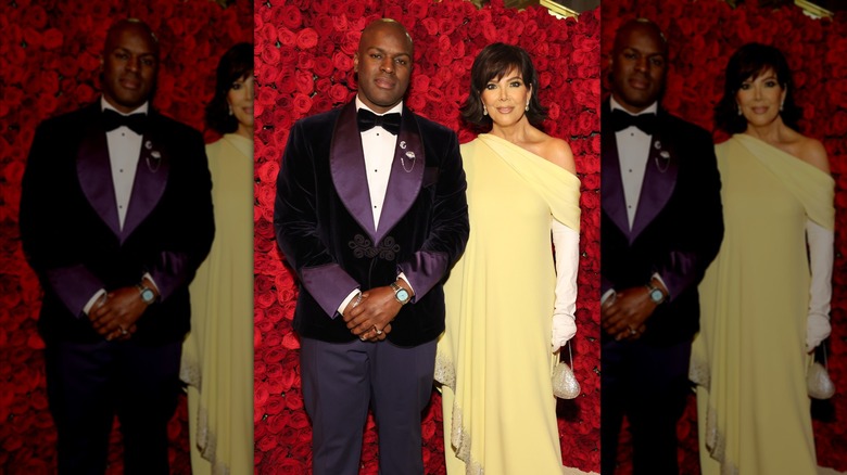 Corey Gamble wears a purple suit and Kris Jenner wears a yellow gown and white gloves as they pose in front of a wall of roses.