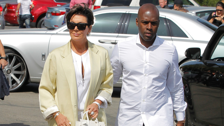 Kris Jenner wears a yellow jacket and walks outdoors beside Corey Gamble, who wears a white shirt.
