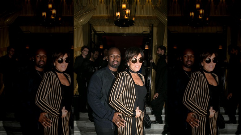 Corey Gamble wearing an all-black outfit and standing behind Kris Jenner, who is wearing a striped suit.