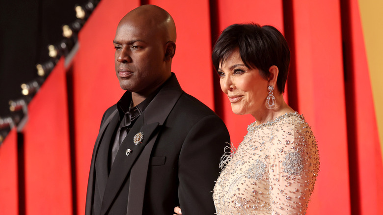 Corey Gamble wearing a black suit and satin shirt and posing beside Kris Jenner, who wears a cream-colored beaded gown.