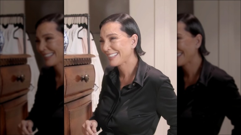 Kris Jenner smiling with her hair slicked down in a black button-up shirt.