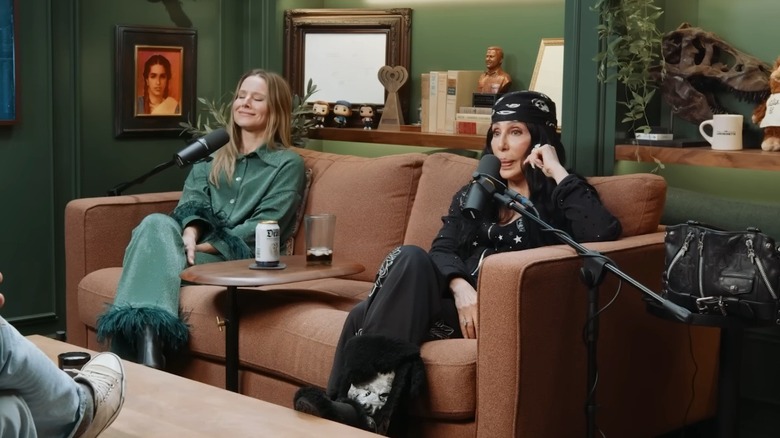 Kristen Bell smiles and closes her eyes while sitting beside Cher on a couch during an "Armchair Expert" episode.