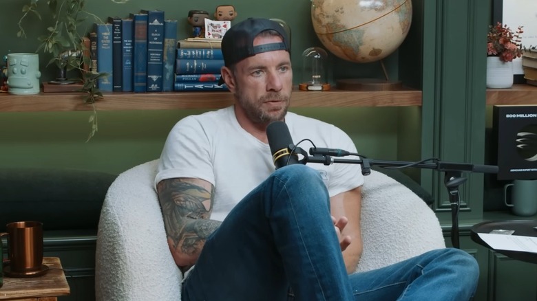 Dax Shepard wearing a backward hat and speaking into a microphone on the set of "Armchair Expert."