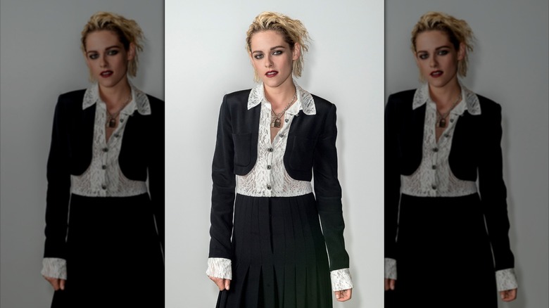 Kristen Stewart with a short, blond bob wearing a black suit against a pale gray backdrop.