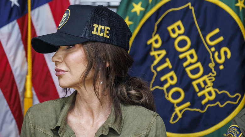 Kristi Noem wearing an ICE hat in front of a border patrol flag and an American flag