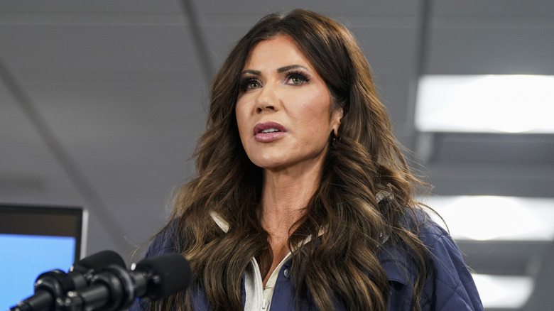 Kristi Noem looking worried in a navy jacket in front of a microphone