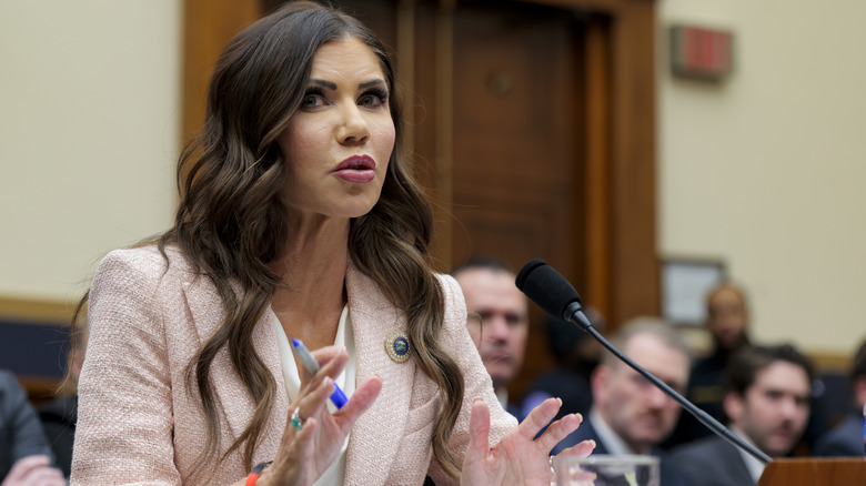 Kristi Noem speaking while gesturing with her hands and holding a pen in a blazer