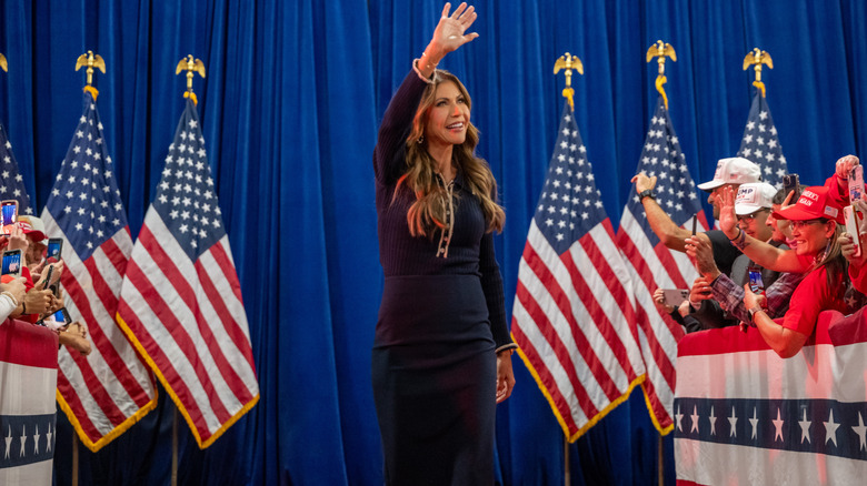 Kristi Noem waving at a crowd in a black dress in front of some American flags