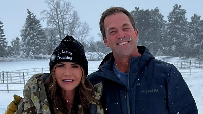 Kristi Noem smiling while wearing a black hat and army jacket next to Bryon Noem who's wearing a coat while it's snowing.