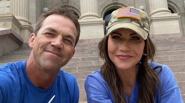Kristi Noem wearing a green cap with an American flag on it smiling next to Bryon Noem who's wearing a blue shirt.