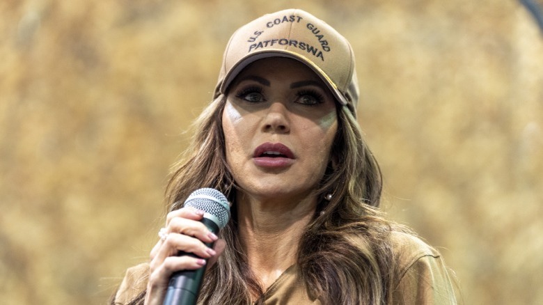 Kristi Noem giving a speech in Bahrain, holding a microphone and wearing a tan Coast Guard hat (2025)
