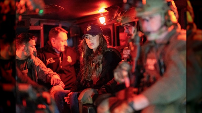 Kristi Noem in a bulletproof vest and cap inside a red lit van, surrounded by people.
