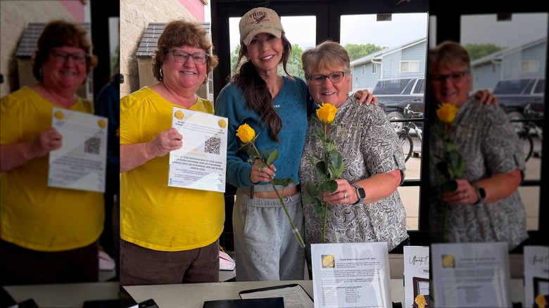 Kristi Noem smiling next to two women while wearing a green long-sleeved shirt, a brown hat, and a pair of sweat pants.
