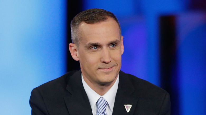 Corey Lewandowski smiling in a suit with his hands clasped on the desk in front of him
