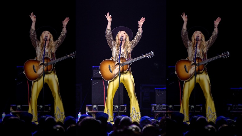 Lainey Wilson performing on stage in yellow pants with her hands in the air.