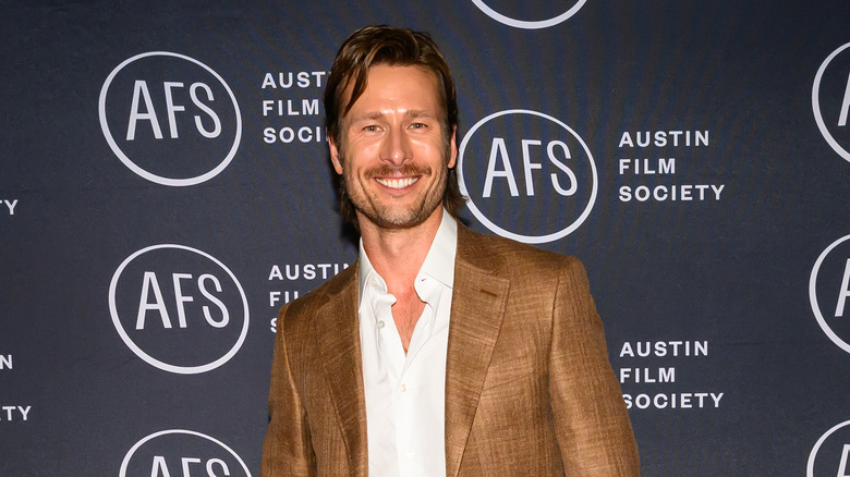 Glen Powell smiles in a brown jacket and white shirt
