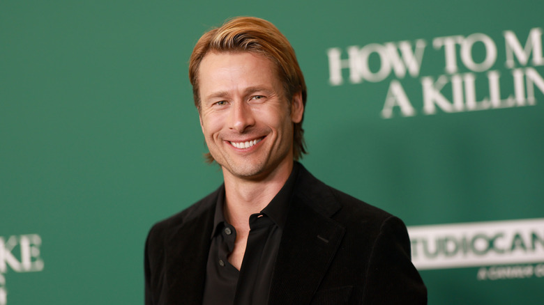 Glen Powell smiles in a black suit and button down shirt