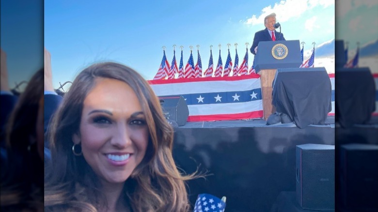 Lauren Boebert smiling while wearing a tan coat and black shirt while President Donald Trump speaks at a podium in the background.