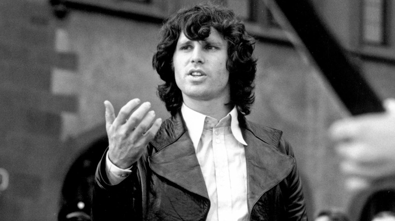 A black-and-white photo of Jim Morrison lifting his right hand in a white shirt and black jacket