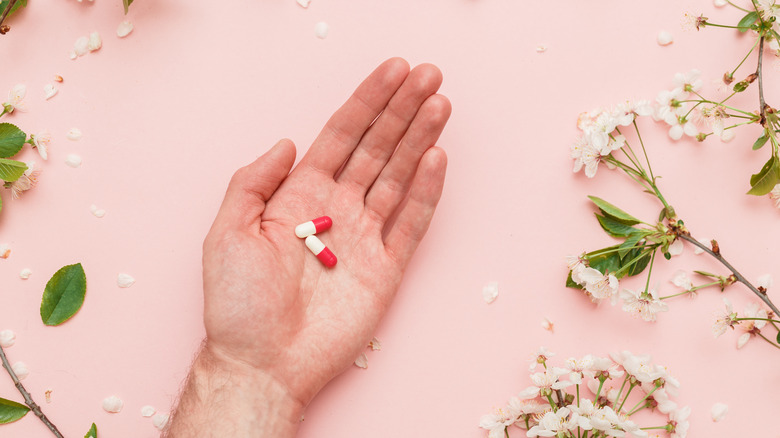 white hand holding two pills with flowers on pink background