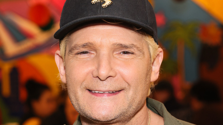 Corey Feldman smiling while posing at an event in a black baseball cap