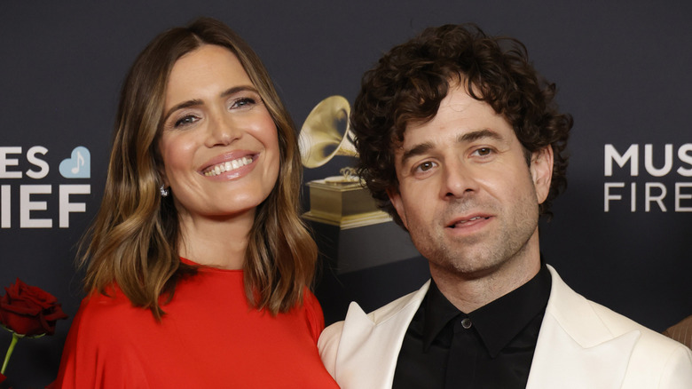 Mandy Moore smiling in a red dress with her husband Taylor Goldsmith in a white suit with a black shirt