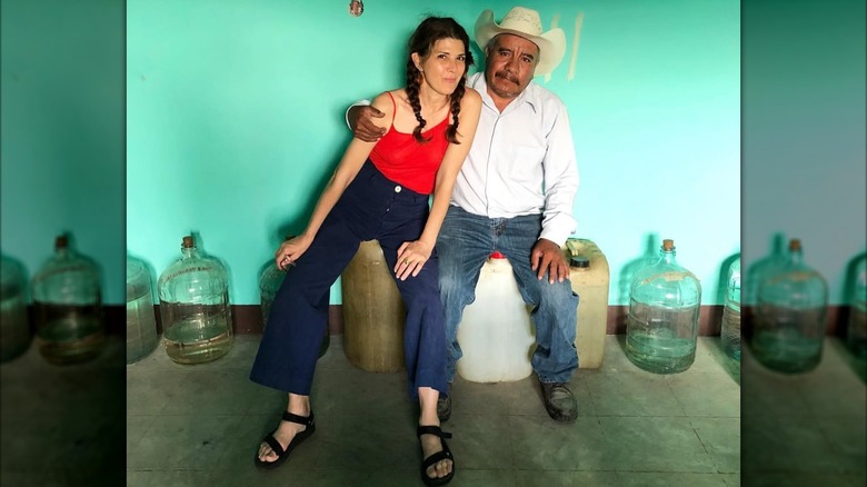 Marisa Tomei wearing her hair in pigtails and seated beside a mezcalero on mezcal containers