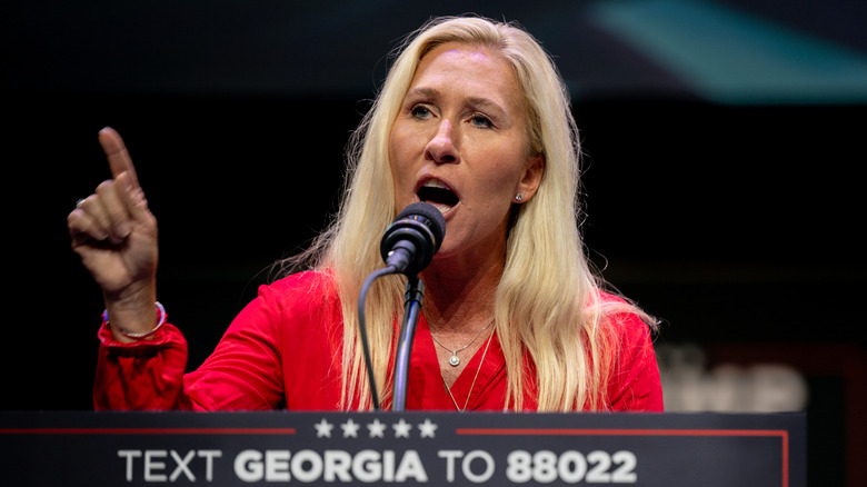 Marjorie Taylor Greene pointing while speaking in front of a podium in a red blouse
