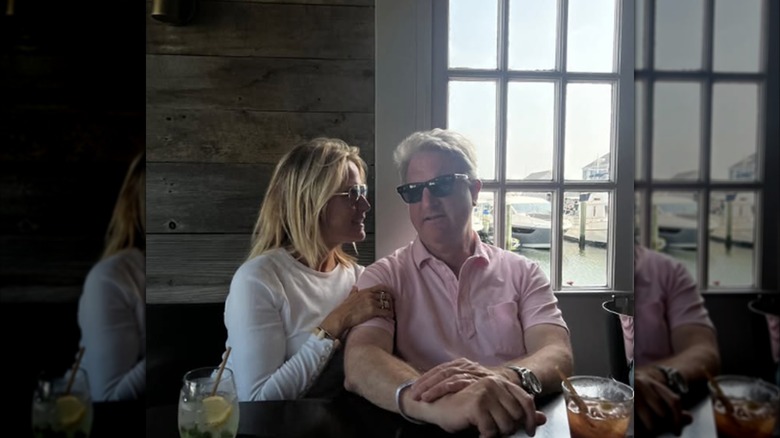 Martha MacCallum wearing a white long-sleeved shirt and sunglasses while smiling next to her husband, Daniel John Gregory, while they're at a restaurant.