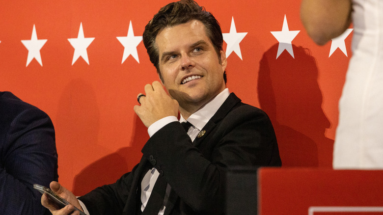 Matt Gaetz smiling in a suit while looking sideways with his phone in his hand