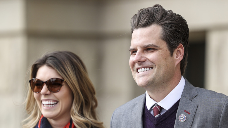 Matt Gaetz smiling in a gray suit and purple sweater next to Ginger Gaetz who's wearing sunglasses and a black coat.