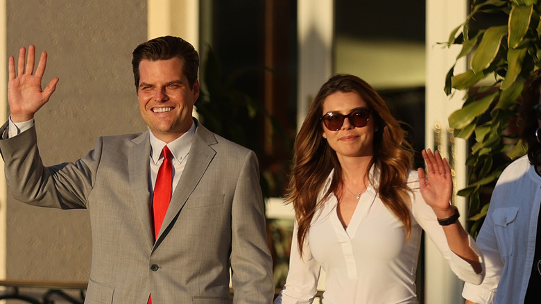 Ginger Gaetz smiling in a white shirt and sunglasses next to her husband Matt Gaetz who's wearing a gray suit and red tie.