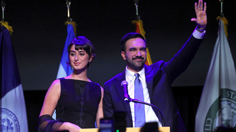 Zohran Mamdani waving while Rama Duwaji stands beside him