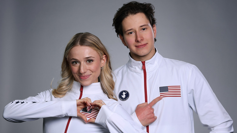 Emelia Zingas and Vadym Kolesnik wear matching white U.S. skating tracksuits and smile while Emelia Zingas makes a heart with her hands