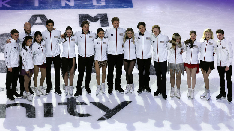 The 2026 U.S. Olympic figure skating team link arms and pose smiling on the ice wearing matching white jackets