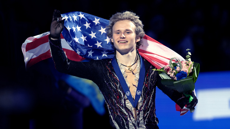 Ilia Malinin smiiles with the American flag behind his back holding a bouquet of flowers in his left hand