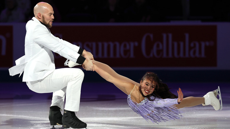 Danny O'Shea wears a white suit and holds Ellie Kam as she smiles and spins in lilac on the ice