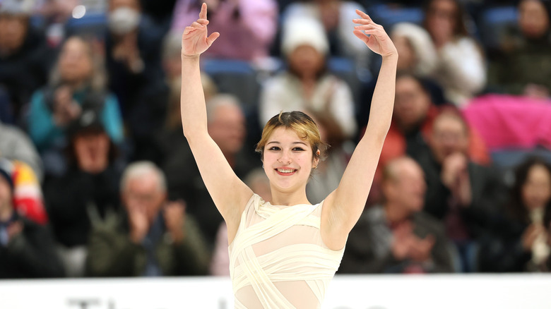 Alysa Liu smiles on the ice with hands in the air in front of a crowd wearing white