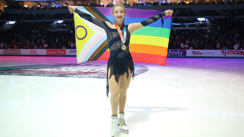 Amber Glenn smiles wearing a medal on ice holding the pride flag behind her