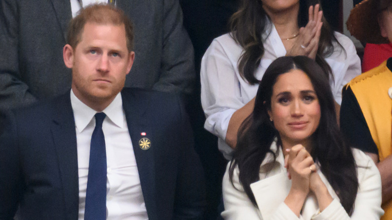Prince Harry in a suit next to Meghan Markle who has her hands together while wearing a white coat.